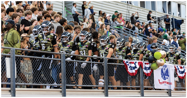 A group of people in football uniforms and helmets stand together on bleachers decorated with patriotic banners and balloons while a crowd watches from behind. Description generated by AI