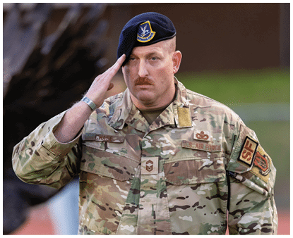 A person in military camouflage uniform and a blue beret is saluting with a serious expression while standing outdoors near a large dark sculpture. Description generated by AI