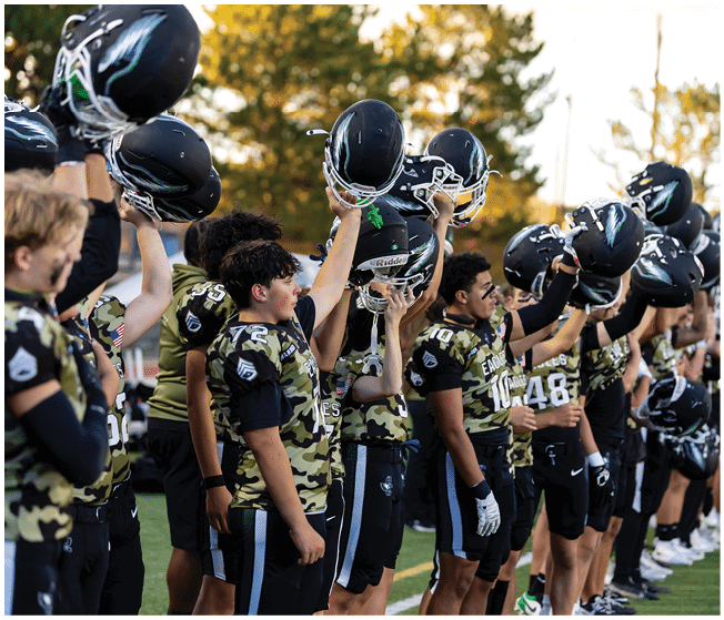 A group of people wearing camouflage sports jerseys and black shorts stand in a line on a field, holding up black helmets with white designs. Description generated by AI