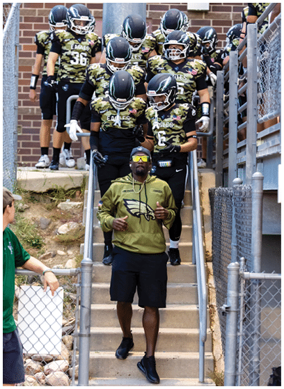 A person wearing sunglasses and a green hoodie with an Eagles logo is standing on stairs in front of a group of people dressed in camouflage football uniforms descending the stairs. Description generated by AI