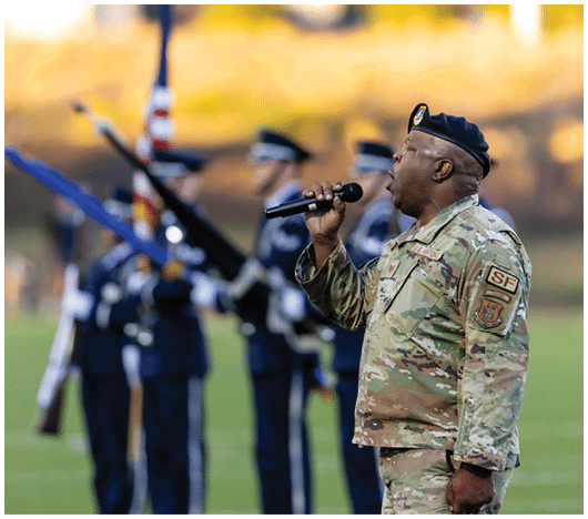 A person in military uniform is singing into a microphone with a color guard holding rifles and flags blurred in the background. Description generated by AI