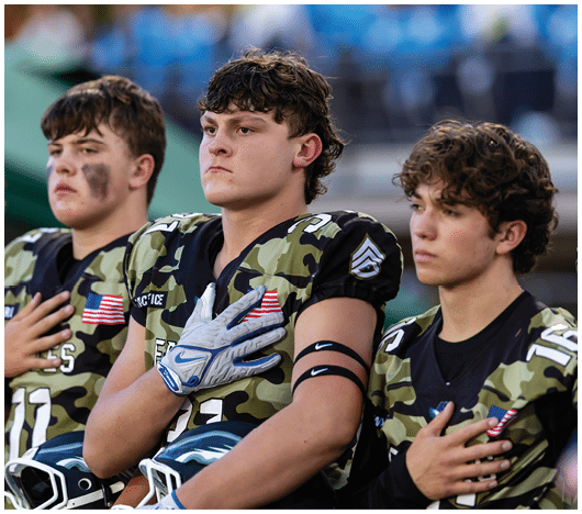 Three people in camouflage football uniforms stand with their hands over their hearts, appearing focused and solemn. Description generated by AI