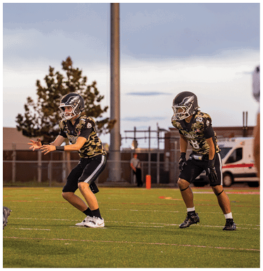 Children wearing football uniforms are playing on a grassy field during a game at dusk with trees and a fence in the background. Description generated by AI