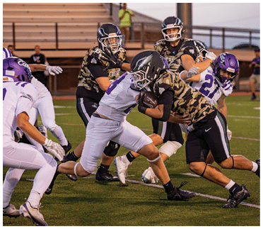People wearing football helmets and uniforms are engaged in a tackle during a game on a grassy field with bleachers in the background. Description generated by AI