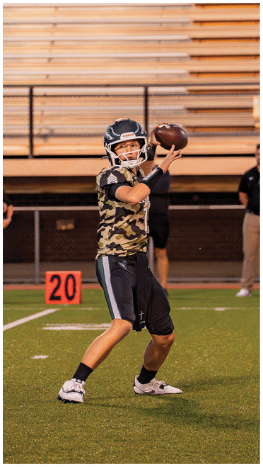 A person wearing a camouflage sports jersey and helmet is preparing to throw a football on a field with empty bleachers and two people standing in the background. Description generated by AI