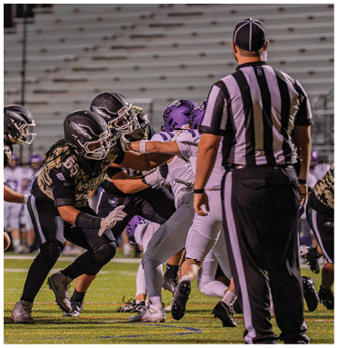 Several people in football uniforms are engaged in a play on a field with a referee standing nearby observing the action. Description generated by AI