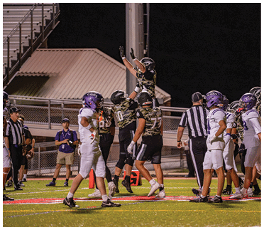 People in football uniforms are celebrating on the field with one person jumping and raising their arms while others look on. Description generated by AI