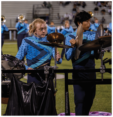 Two people in matching blue and black uniforms play percussion instruments on a field with a marching band in the background. Description generated by AI