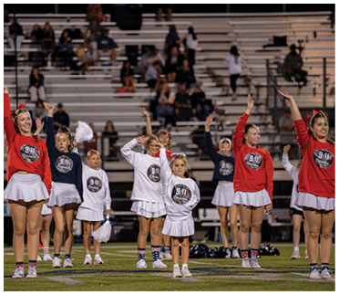 A group of people wearing matching cheerleading outfits with 9/11 remembrance logos are standing on a field with their arms raised, with bleachers and spectators in the background. Description generated by AI