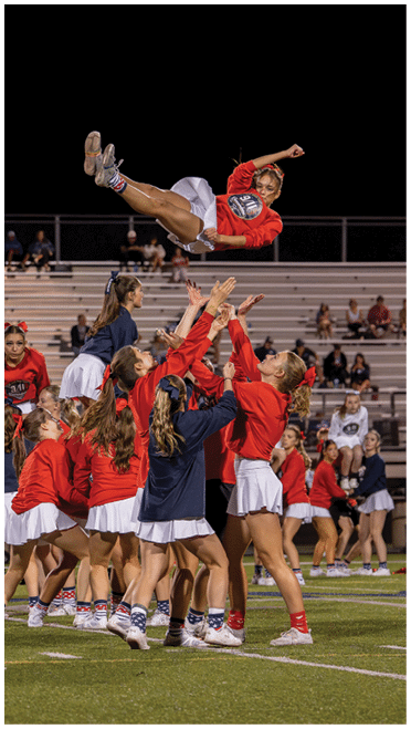A group of people wearing red and navy tops with white skirts are performing a cheerleading stunt on a sports field at night with one person mid-air above the others. Description generated by AI