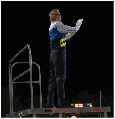 A person wearing a blue and white top with dark pants stands on a raised platform at night with a dark sky and some distant lights in the background. Description generated by AI