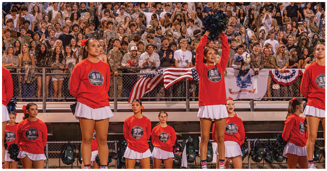 People wearing matching red sweatshirts and white skirts perform a cheerleading routine in front of a large crowd at a stadium with American flags and patriotic decorations. Description generated by AI