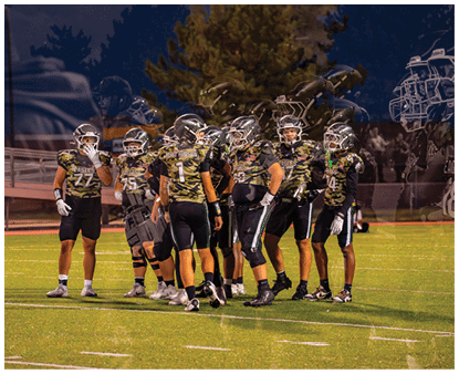 A group of people wearing camouflage-patterned football uniforms and helmets are standing together on a lit football field at night. Description generated by AI