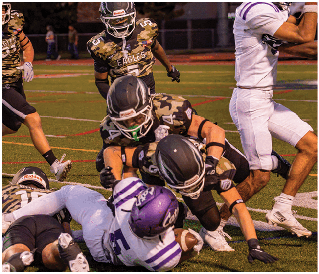 Several football players in camouflage and white uniforms are engaged in a tackle on a grassy field during a game. Description generated by AI