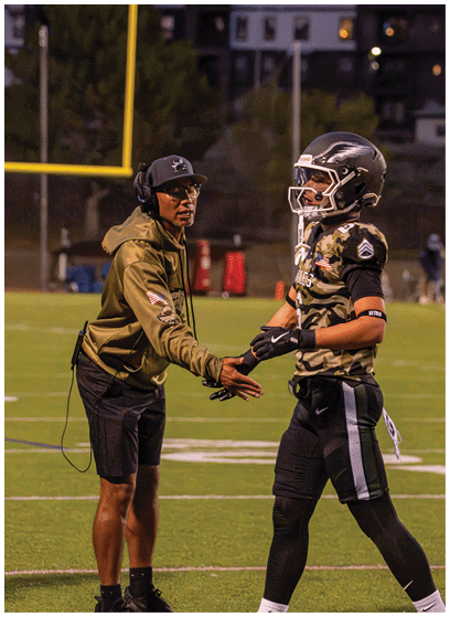 An adult wearing a headset and casual sportswear is shaking hands with a person in a football uniform and helmet on a football field at dusk. Description generated by AI