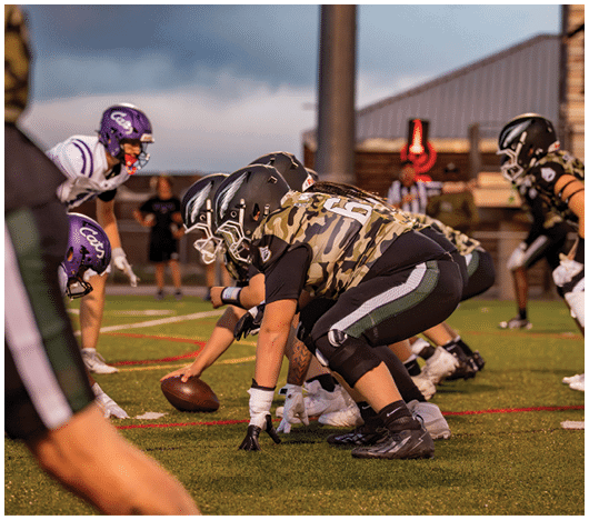 People in football uniforms are lined up on a field facing each other, preparing for a play during a game. Description generated by AI