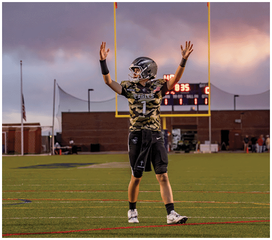 A person wearing a camouflage sports jersey and helmet stands on a football field with arms raised near the goalposts and a scoreboard in the background. Description generated by AI