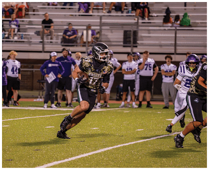 A person wearing a camouflage football jersey with the word Eagles is running on a field holding a football while other players and coaches stand in the background near bleachers. Description generated by AI