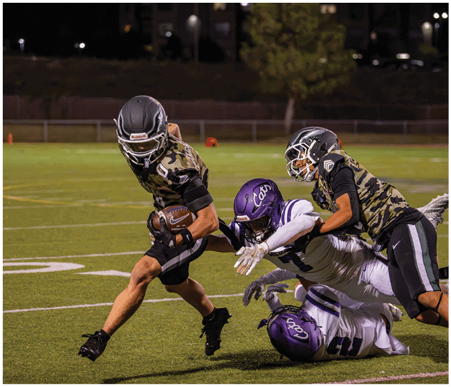 A person in a camouflage sports uniform is running with a football while two other people in similar uniforms tackle a person in a white and purple football uniform on a grassy field at night. Description generated by AI