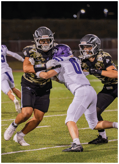 Two people in football uniforms are engaged in a tackle on a grassy field while another player runs in the background. Description generated by AI