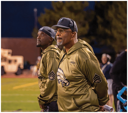 Two adults wearing matching green hooded jackets with eagle logos stand side by side on a sports field at night, one holding a rolled-up paper. Description generated by AI