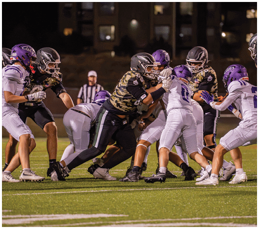 Adults wearing football helmets and uniforms are engaged in a play on a well-lit field at night with a referee in the background. Description generated by AI