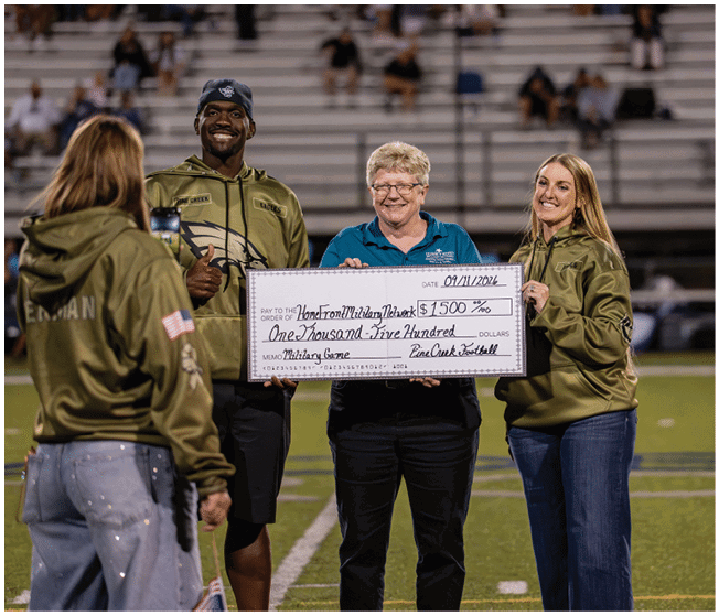 Three adults smiling and holding a large ceremonial check on a sports field with bleachers and spectators in the background. Description generated by AI