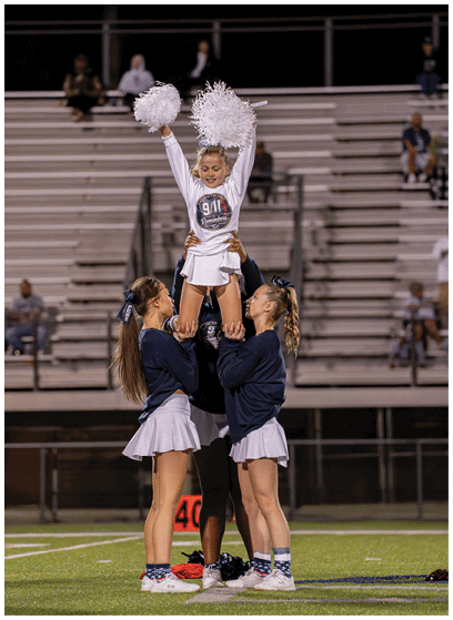 Four people in cheerleading uniforms perform a stunt on a sports field with empty bleachers in the background. Description generated by AI