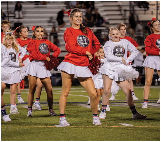 A group of adults and children wearing matching cheerleading outfits with red and white tops and white skirts are performing a cheer routine on a sports field. Description generated by AI