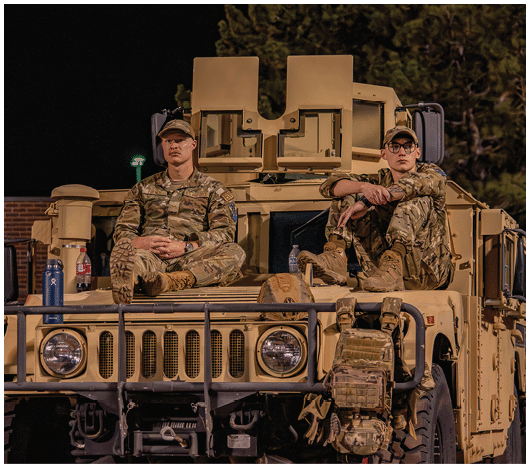 Two people in military camouflage uniforms sit on the hood of a tan armored vehicle at night. Description generated by AI