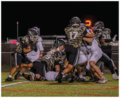 Several football players in camouflage jerseys and helmets are engaged in a tackle on a grassy field under stadium lights at night. Description generated by AI