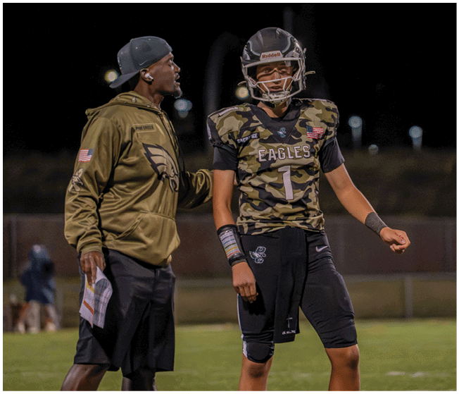 A person wearing a sports hoodie and cap is talking to a person in a football uniform and helmet on a field at night. Description generated by AI
