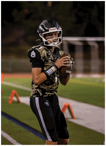 A person wearing a camouflage sports jersey and helmet is holding a football and preparing to throw it on a lit outdoor field at night. Description generated by AI