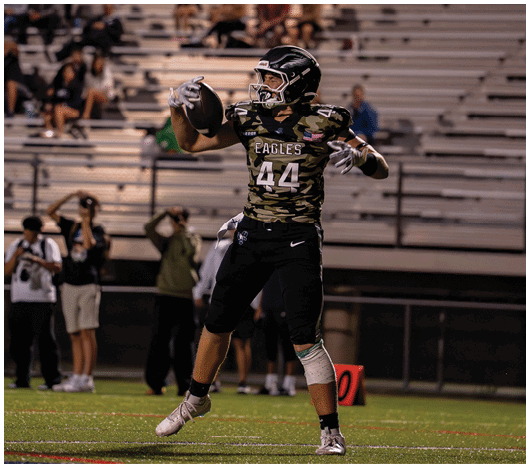 A person wearing a camouflage football jersey with the number 44 is holding a football and preparing to throw it on a field with empty bleachers in the background. Description generated by AI