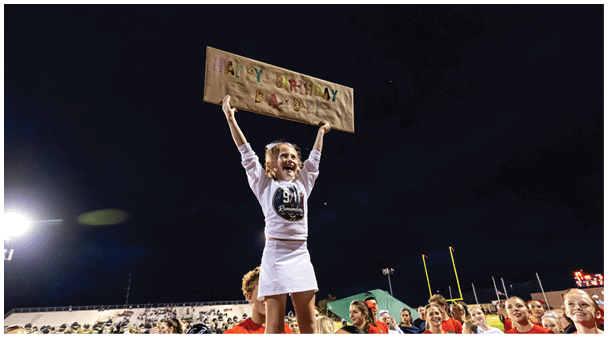 People wearing matching red sweatshirts and white skirts perform a cheerleading routine in front of a large crowd at a stadium with American flags and patriotic decorations. Description generated by AI