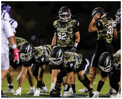 A group of people wearing camouflage-patterned football uniforms and helmets are standing together on a lit football field at night. Description generated by AI