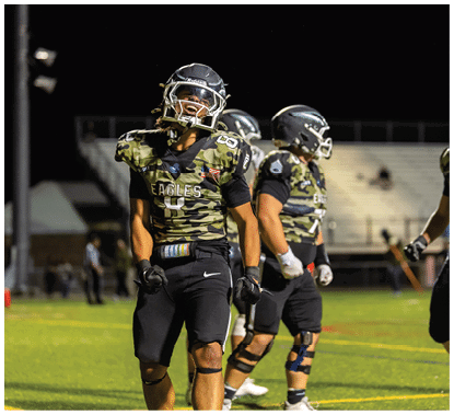 A group of people wearing camouflage-patterned football uniforms and helmets are standing together on a lit football field at night. Description generated by AI