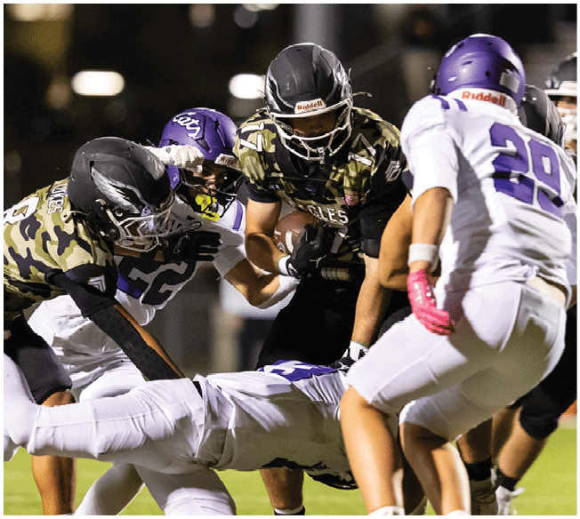 Several football players in camouflage and white uniforms are engaged in a tackle on a grassy field during a game. Description generated by AI