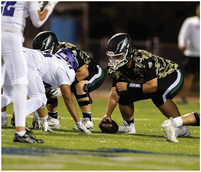 Several football players in camouflage and white uniforms are engaged in a tackle on a grassy field during a game. Description generated by AI
