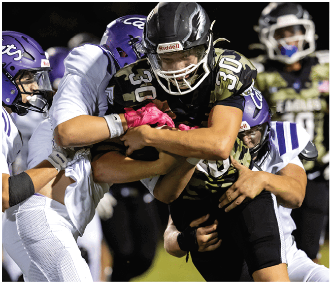 Several football players in camouflage and white uniforms are engaged in a tackle on a grassy field during a game. Description generated by AI