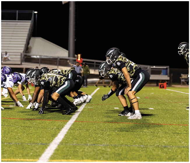 Several football players in camouflage and white uniforms are engaged in a tackle on a grassy field during a game. Description generated by AI