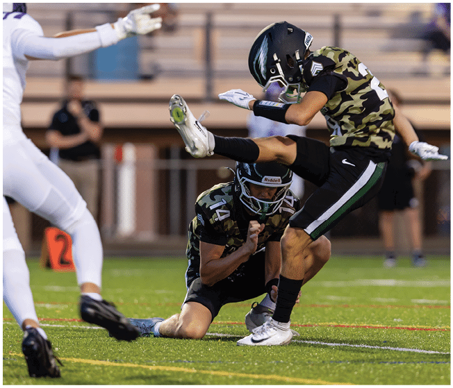 Several football players in camouflage and white uniforms are engaged in a tackle on a grassy field during a game. Description generated by AI
