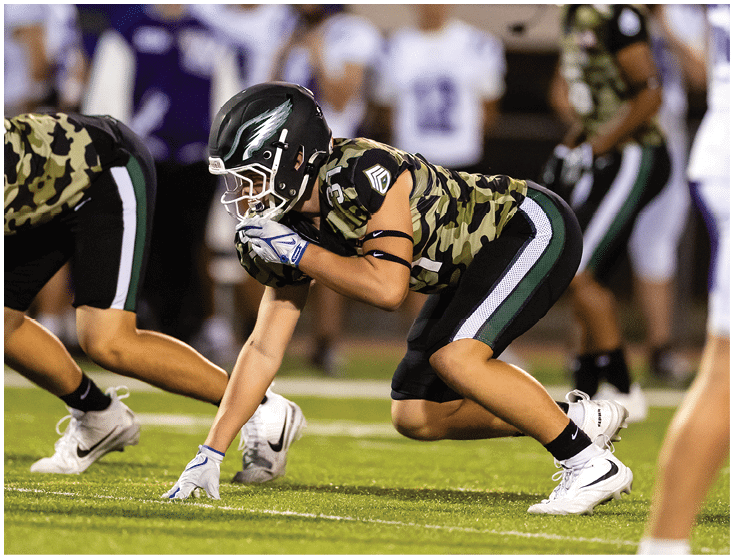 Several football players in camouflage and white uniforms are engaged in a tackle on a grassy field during a game. Description generated by AI