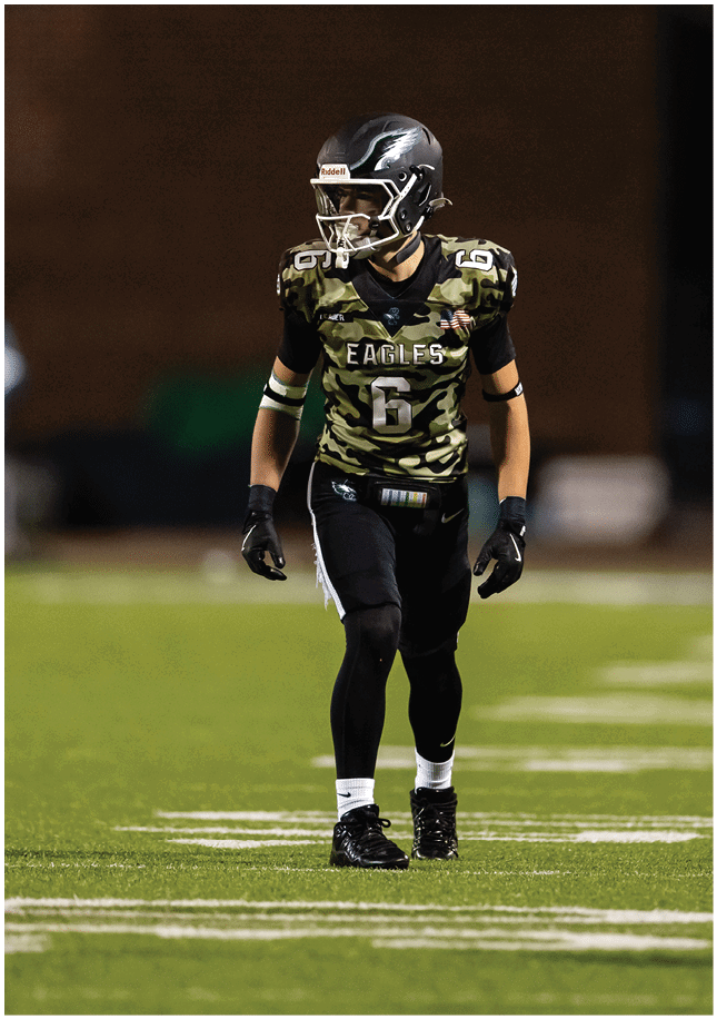 An adult wearing a headset and casual sportswear is shaking hands with a person in a football uniform and helmet on a football field at dusk. Description generated by AI