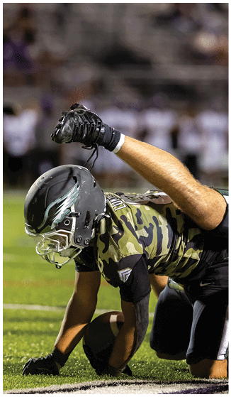 An adult wearing a headset and casual sportswear is shaking hands with a person in a football uniform and helmet on a football field at dusk. Description generated by AI