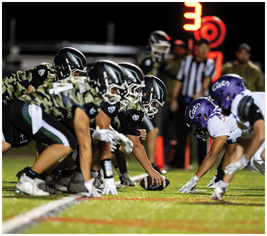 People in football uniforms are lined up on a field facing each other, preparing for a play during a game. Description generated by AI