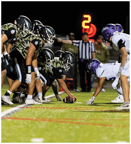 A person wearing a camouflage sports jersey and helmet stands on a football field with arms raised near the goalposts and a scoreboard in the background. Description generated by AI