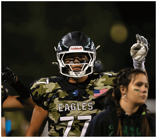 A person wearing a camouflage sports jersey and helmet stands on a football field with arms raised near the goalposts and a scoreboard in the background. Description generated by AI