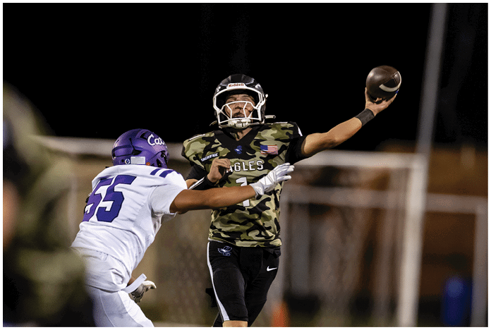 A person wearing a camouflage sports jersey and helmet stands on a football field with arms raised near the goalposts and a scoreboard in the background. Description generated by AI