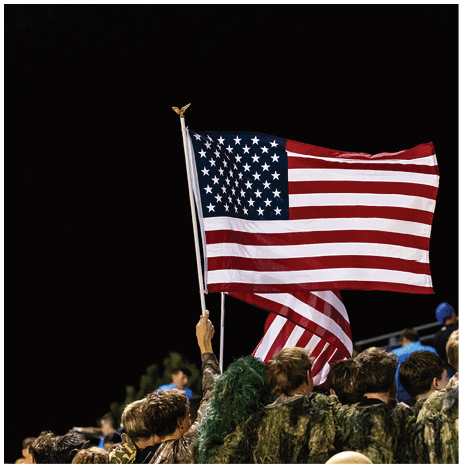A person wearing a camouflage sports jersey and helmet stands on a football field with arms raised near the goalposts and a scoreboard in the background. Description generated by AI